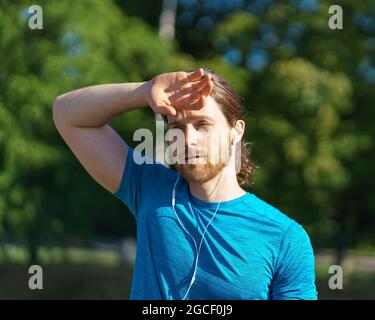 Stanco sfinito giovane uomo sportivo strofinando sudore dalla fronte mentre si esercita all'aperto Foto Stock