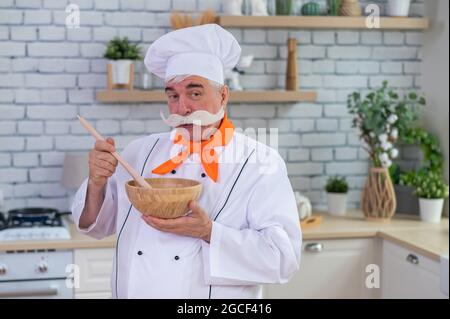 Un uomo anziano con baffi grigi nel cappello di uno chef sta cucinando in cucina. Foto Stock