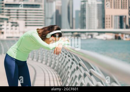 Donna che soffre di respiro corto o asma si appoggia contro una recinzione e riposa dopo un allenamento in corsa sull'argine di Dubai Marina Foto Stock