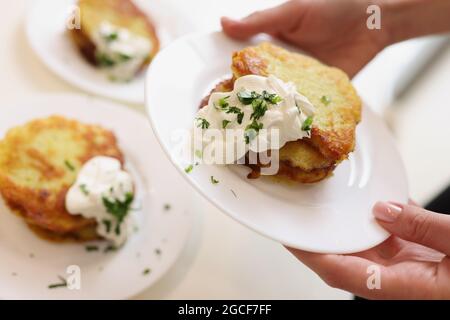 La donna tiene in mano un piatto con frittelle di patate e panna acida ed erbe aromatiche Foto Stock