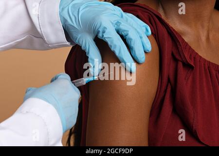 Cropped di mani di infermiera in guanti medici protettivi che fanno l'iniezione intramuscolare nella spalla per donna nera irriconoscibile, foto di closeup. Vaccinat Foto Stock