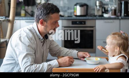 Un uomo attento con la barba nutre la sua figlia piccola. Una bambina si siede a un tavolo da pranzo per bambini e cena con suo padre. Felice paternità. Foto Stock