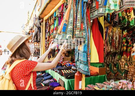 Una donna in una maschera medica a causa della pandemia del coronavirus sceglie i tessuti ed i vestiti etnici ad un bazar locale o ad un mercato in Africa Foto Stock