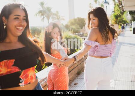 Tre ragazze latina che passeggiano sorridendo per la strada godendo la loro amicizia. Giovani donne che camminano sul fiume ridendo. Ragazze che trascorrono una giornata di sole Foto Stock