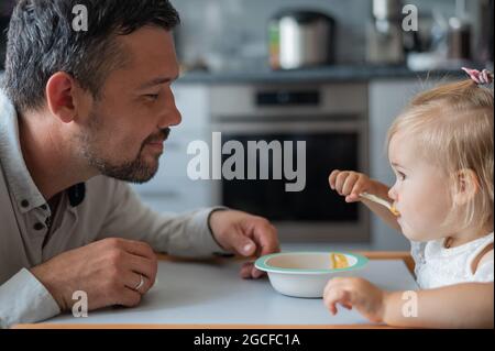 Un uomo attento con la barba nutre la sua figlia piccola. Una bambina si siede a un tavolo da pranzo per bambini e cena con suo padre. Felice paternità. Foto Stock