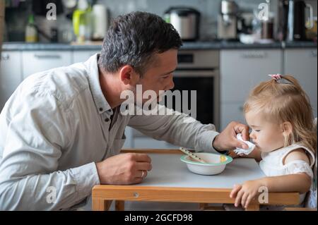 Un uomo attento con la barba nutre la sua figlia piccola. Una bambina si siede a un tavolo da pranzo per bambini e cena con suo padre. Felice paternità. Foto Stock