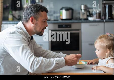 Un uomo attento con la barba nutre la sua figlia piccola. Una bambina si siede a un tavolo da pranzo per bambini e cena con suo padre. Felice paternità. Foto Stock