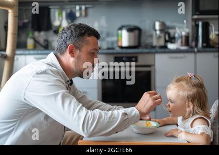 Un uomo attento con la barba nutre la sua figlia piccola. Una bambina si siede a un tavolo da pranzo per bambini e cena con suo padre. Felice paternità. Foto Stock