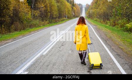 Una donna dai capelli rossi in un cappotto giallo cammina con una valigia gialla lungo l'autostrada in autunno. Foto Stock