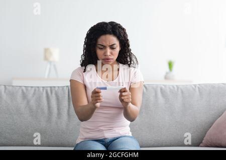 Cattive notizie, infertilità, gravidanza non pianificata, ginecologia e problemi di salute femminile. Triste infelice giovane donna afroamericana sul divano guardando la preparazione Foto Stock