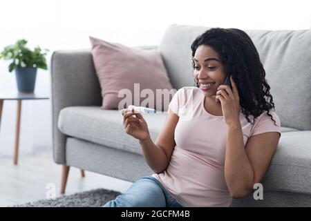 Ginecologia, salute femminile, attesa del bambino e grandi notizie. Sorridente donna afroamericana millenaria sul divano guardando il test di gravidanza e di chiamata Foto Stock