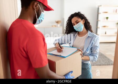 Concetto di consegna sicura, posta, persone e spedizione. Giovane donna in maschera facciale che conferma l'ordine a casa, firma modulo cartaceo dopo aver ricevuto il pacco da bl Foto Stock