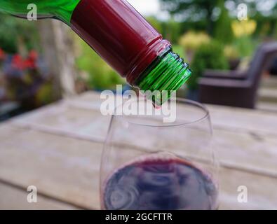 versando l'ultima goccia di vino rosso dalla bottiglia in un bicchiere, un tavolo di legno e uno sfondo di giardino sfocato Foto Stock