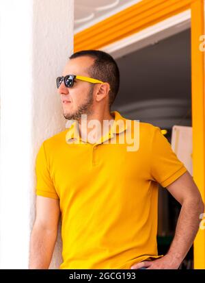 ripresa verticale di un giovane uomo con maglietta gialla e occhiali da sole in piedi vicino a un muro. Foto Stock