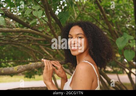 Moda primo piano ritratto di ridere attraente giovane naturalmente bella donna afroamericana con capelli afro e denti perfetti sorridono in natura parco in verde fogliame Foto Stock