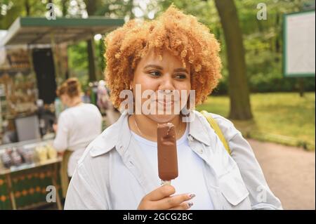 Stile di vita ritratto di sorridente giovane naturalmente bella più taglia Africana americana donna con capelli rossi afro mangiare gelato cioccolato popsicle nel parco pubblico. Diversità e concetto positivo per il corpo Foto Stock