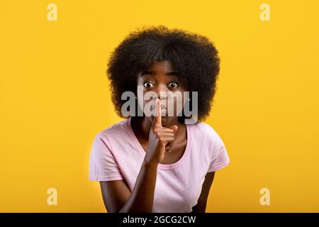 Mantenere il silenzio. Signora nera che mostra il gesto shh alla macchina fotografica, stanfing sopra sfondo giallo, scatto studio. Giovane donna afroamericana che tiene il dito vicino Foto Stock