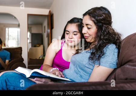 Ragazza ispanica adolescente con sindrome di Down e la sua mamma che tiene un libro a casa, in concetto di disabilità in America Latina Foto Stock