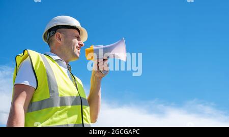 Il costruttore in un casco bianco urla in un megafono contro uno sfondo blu del cielo Foto Stock