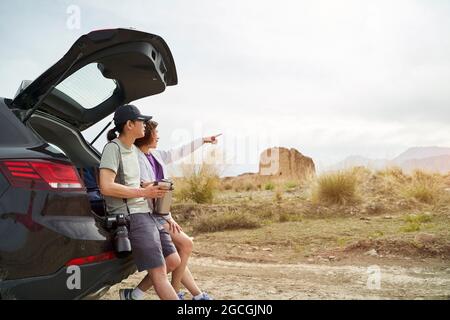 la coppia asiatica si appoggia contro il retro dell'auto guardando la vista mentre si beve caffè in un sito storico desolato Foto Stock