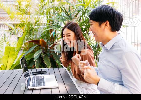 Felice giovane asiatico uomo e donna si sentono entusiasti di ricevere buone notizie mentre navigano il computer portatile e fanno videochiamata in un moderno bar Foto Stock