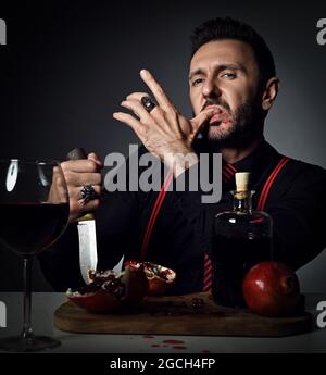 Uomo insolente in camicia nera e sospenditori a tavola con melograno fresco e succo, tiene il coltello e leccò il dito Foto Stock