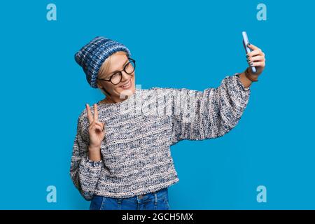 Immagine elegante con donna in maglia cappello che fa selfie. Look alla moda. Gesto di pace. Foto Stock