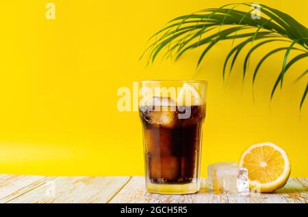 Un bicchiere di bevanda analcolica fredda con limone su pavimento in legno con foglia di cocco e sfondo giallo. Concetto di bevanda estiva. Foto Stock