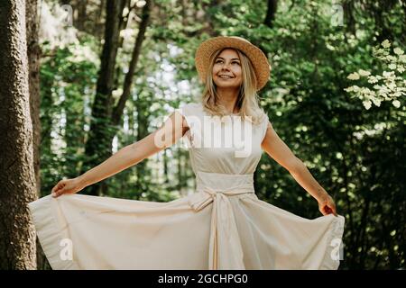 Ritratto esterno di giovane donna in cappello di paglia che tiene l'orlo del suo vestito bianco mentre cammina nel parco o nella foresta Foto Stock