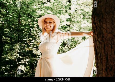 Ritratto esterno di giovane donna in cappello di paglia che tiene l'orlo del suo vestito bianco mentre cammina nel parco o nella foresta Foto Stock