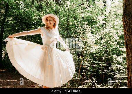 Ritratto esterno di giovane donna in cappello di paglia che tiene l'orlo del suo vestito bianco mentre cammina nel parco o nella foresta Foto Stock