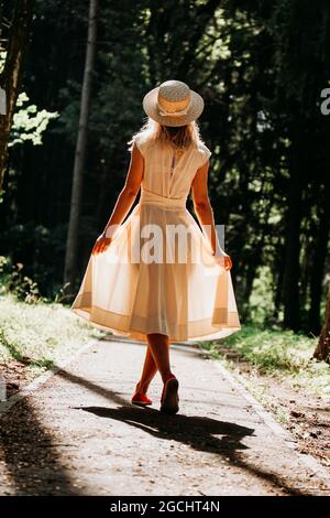 Una giovane donna in un vestito bianco e un cappello di paglia cammina attraverso i boschi. Fata foresta. Vista posteriore Foto Stock