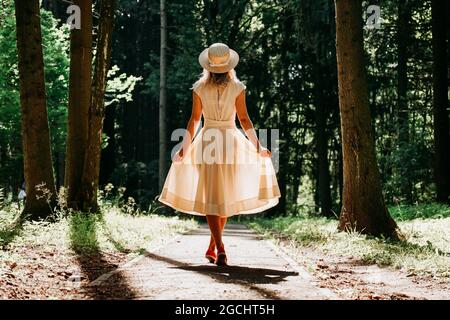 Una giovane donna in un vestito bianco e un cappello di paglia cammina attraverso i boschi. Fata foresta. Vista posteriore Foto Stock