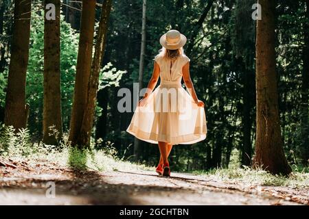 Una giovane donna in un vestito bianco e un cappello di paglia cammina attraverso i boschi. Fata foresta. Vista posteriore Foto Stock