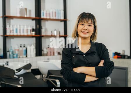 Ritratto di felice successo giovane proprietario di salone asiatico in piedi indossando grembiule e occhiali con le mani ripiegate sorridendo mentre si guarda la macchina fotografica Foto Stock