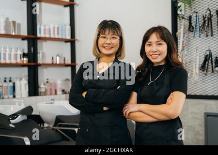 Ritratto di squadra di felice successo giovane asiatico hairstylist in piedi con grembiule e occhiali con le mani ripiegate sorridendo mentre si guarda la macchina fotografica Foto Stock