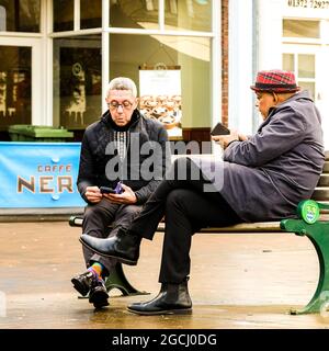 Dicembre 2020, Epsom Surrey, Londra Regno Unito, due uomini anziani amici seduta panca che parla controllare messaggi telefonici Foto Stock