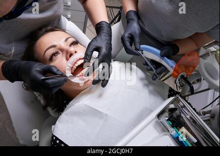 Bella donna in sedia dentale durante la procedura di installazione dei rinforzi ai denti superiore e inferiore. Dentista e assistente che lavorano insieme, strumenti dentali nelle loro mani. Vista dall'alto. Concetto di odontoiatria Foto Stock