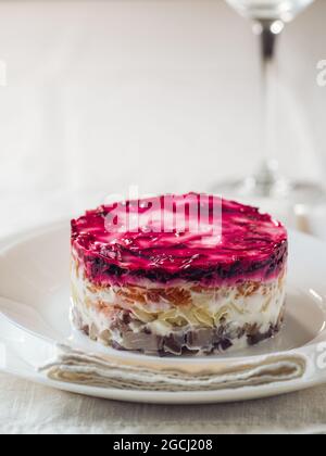 Aringa con insalata a strati sotto il cappotto di pelliccia sul tavolo. Porzione di insalata tradizionale russa con aringhe, barbabietole e altre verdure su tovaglia di lino bianco. Spazio di copia. Verticale. Foto Stock
