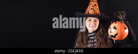 Bandiera della gente di Halloween. Una donna in un costume di strega su uno sfondo nero tiene la zucca secchio con caramella e sorriso. Posizione per il testo dello spazio di copia. Foto Stock