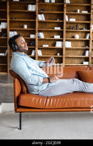 Tempo di pausa. Un ragazzo indiano soddisfatto prende una pausa e riposandosi sul divano e ascoltando musica, uomo ispanico calmo e sorridente in cuffie godendo audiolibri o brani preferiti con gli occhi chiusi Foto Stock