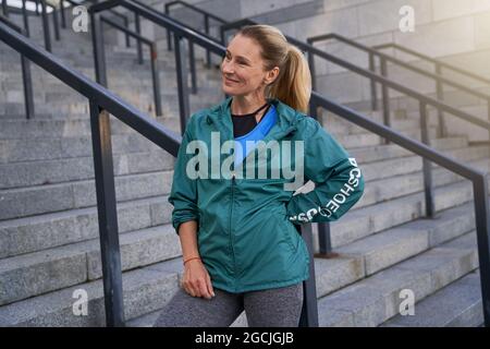 Allegra sportivo di mezza età in abbigliamento sportivo sorridente in piedi fuori dopo l'allenamento in città Foto Stock