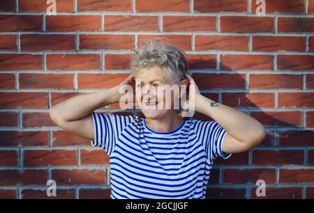 Donna anziana in piedi all'aperto contro muro di mattoni sfondo, rilassante. Foto Stock