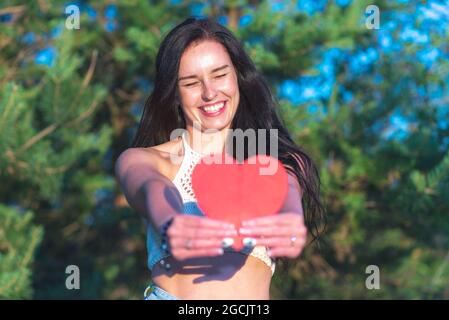 Bella brunetta che mostra rosso cuore cartolina sorridente natura sognante estate soleggiato day.Woman in natura tenendo un cuore di carta rosso in front.Green sfondo. Foto Stock