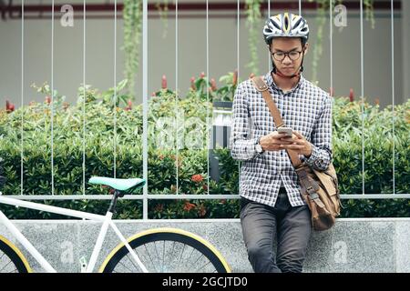 Un giovane studente universitario serio che si appoggia sul recinto accanto alla sua bicicletta e che mette in SMS amici o giochi su smartphone Foto Stock