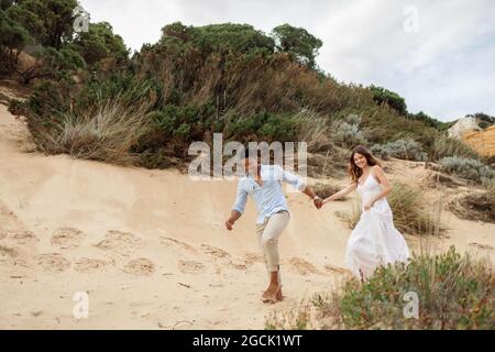 Felice ponte multirazziale e sposo tenendo le mani e correndo lungo la collina di sabbia il giorno di nozze in natura Foto Stock