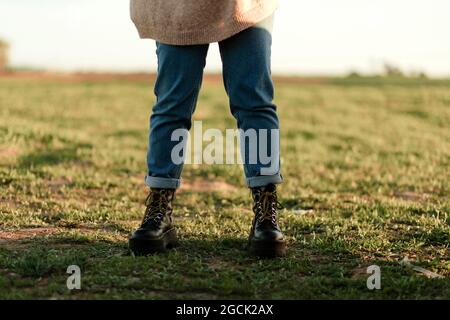 Crop anonimo viaggiatore femminile che indossa maglia pullover con jeans e stivali in pelle nera in piedi in verde campo erboso in campagna primavera Foto Stock