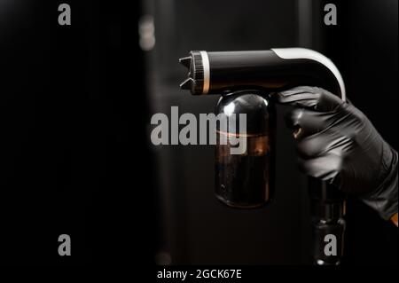 Primo piano dell'apparecchiatura di abbronzatura istantanea nelle mani di un maestro. Vernice spray a pistola per pelle su sfondo nero Foto Stock