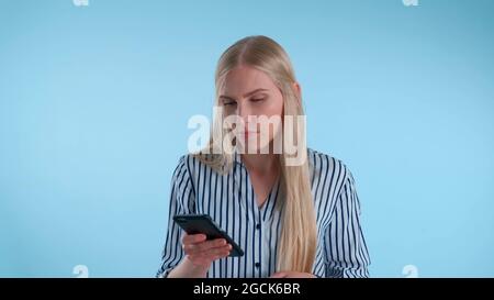 Giovane donna bionda che tiene il mento con la mano mentre pensa a qualcosa su sfondo blu. Guarda lo smartphone e pianifica. Foto Stock