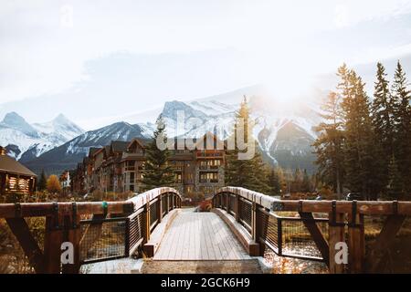 Pittoresco paesaggio autunnale con ponte e case di legno situato vicino al fiume contro le montagne innevate nella città di Canmore vicino al Parco Nazionale di Banff Foto Stock